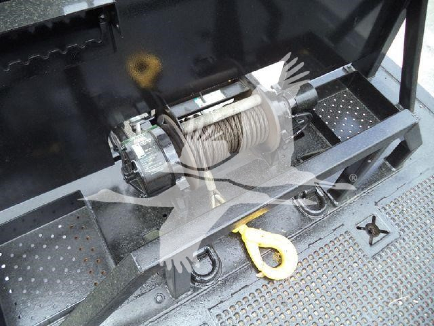 Truck equipment bay with mounted electric winch and steel cable, eyelet hook, and step plate. Visible are coil winch drum, motor, and mounting frame.