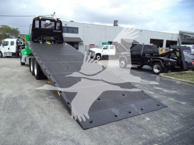 Peterbilt 348 rollback truck with a lowered flatbed ramp, side rails, and a black cab; other trucks in the background.
