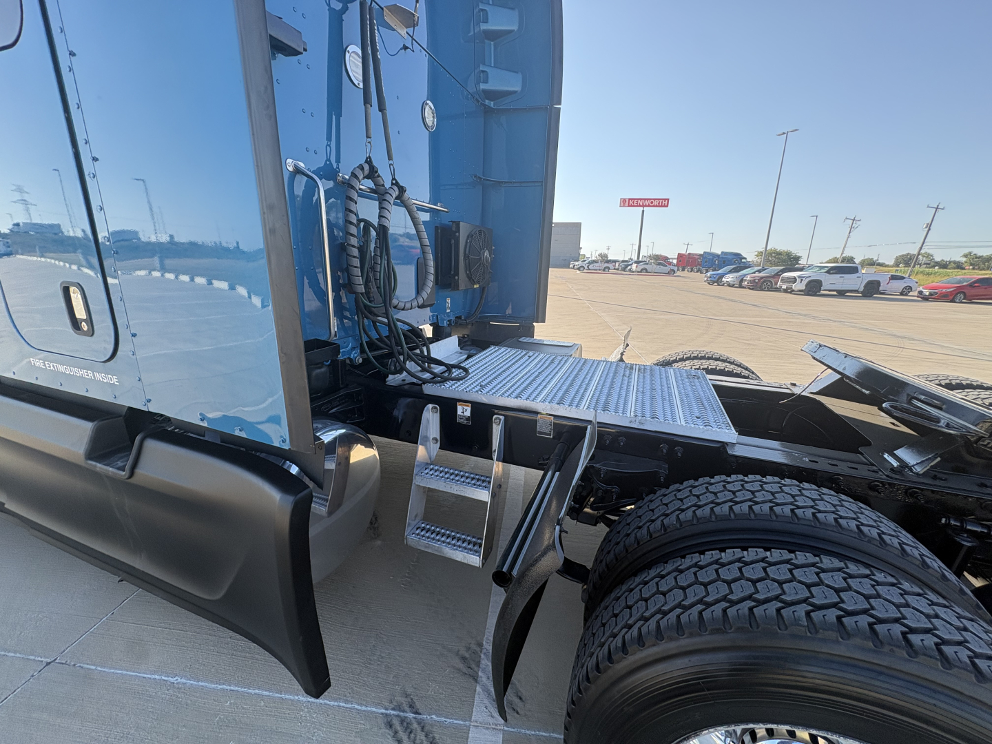 Blue Peterbilt 579 cab rear/side with chrome steps and dual rear wheels, visible metal platform and steps, sleeper tractor unit, parking lot in background.