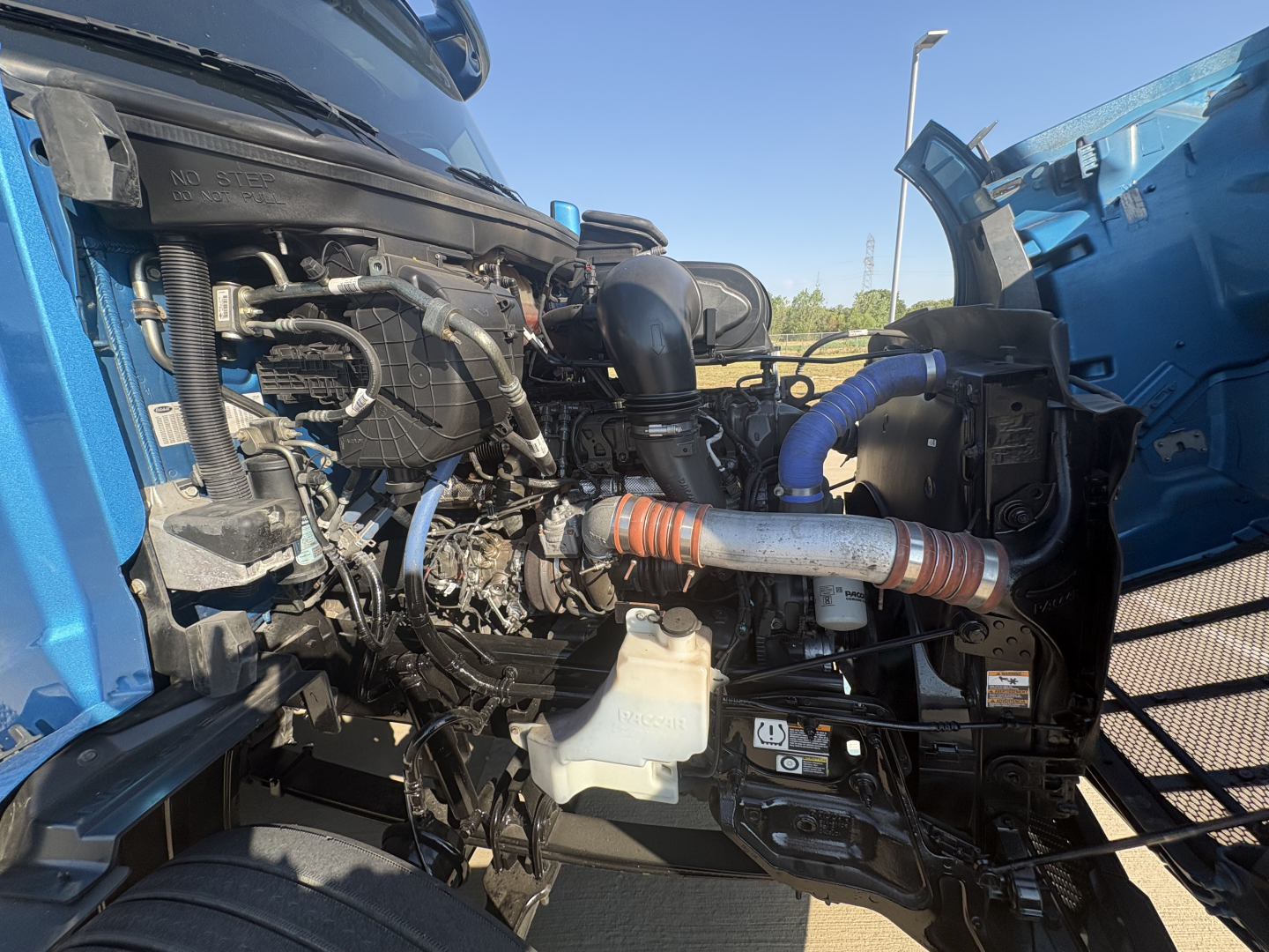 Peterbilt 579 engine bay view with components such as intake, hoses, and coolant reservoir visible in a blue truck with hood open.