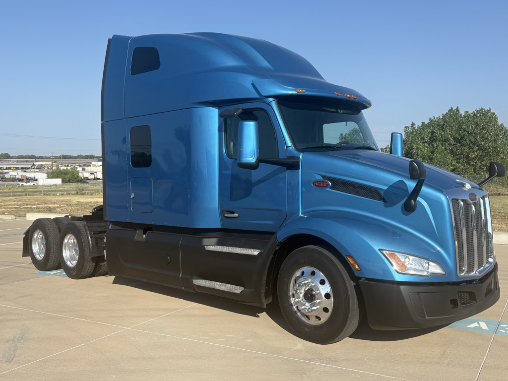 Blue 2022 Peterbilt 579 conventional sleeper truck captured from the passenger side showing front and cab with three axles of rear bogie and chrome wheels.