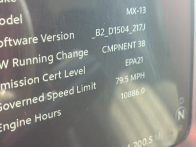 Truck instrument cluster screen showing model MX-13 software version and settings text; includes labels such as Engine Hours, Governed Speed Limit, and MPH reading, with a reflection glare.