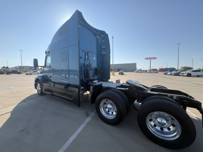 Peterbilt 579 tractor cab from rear-left angle, with dual rear axle and fifth wheel, blue exterior, chrome wheels, highway parking lot background.