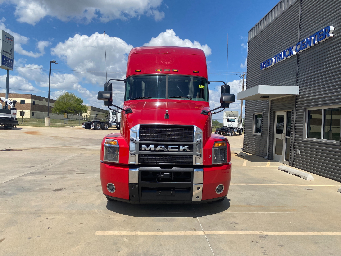 Red Mack AN64T70 conventional sleeper truck facing camera in lot with dealership building on right.