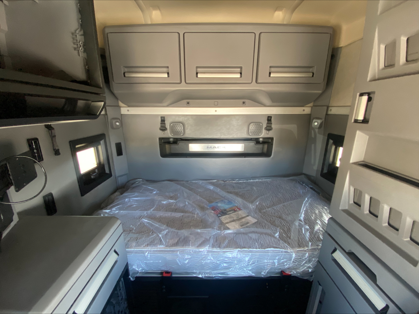 Mack AN64T70 sleeper cab interior showing sleeper bed with plastic cover, overhead cabinets, and side storage compartments.