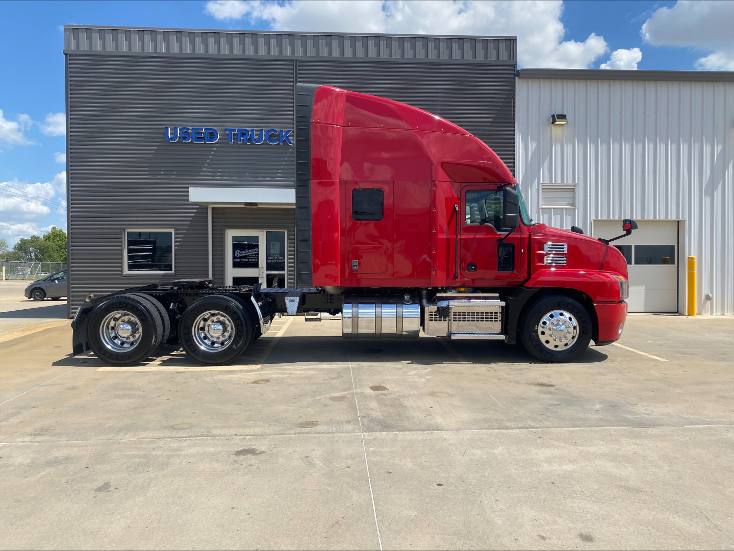 Mack AN64T70 sleeper cab tractor in red, side view, with chrome fuel tanks and triple rear axles, in front of a used truck building.
