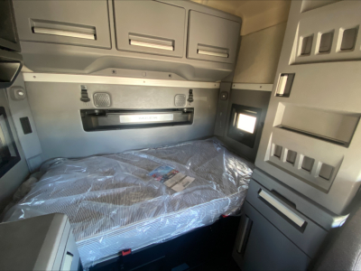 Truck sleeper cabin interior with grey cabinetry and a packaged mattress on the bunk. Upper cabinets and shelves along the walls; small window on the side.