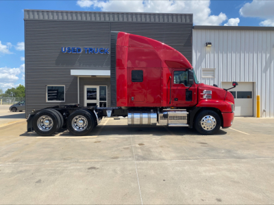 Mack AN64T70 sleeper cab tractor in red, side view, with chrome fuel tanks and triple rear axles, in front of a used truck building.