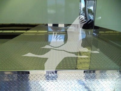 Diamond plate cargo deck and rear step of a Mack MD6 rollback truck showing treadplate surface and reflection in cab window.
