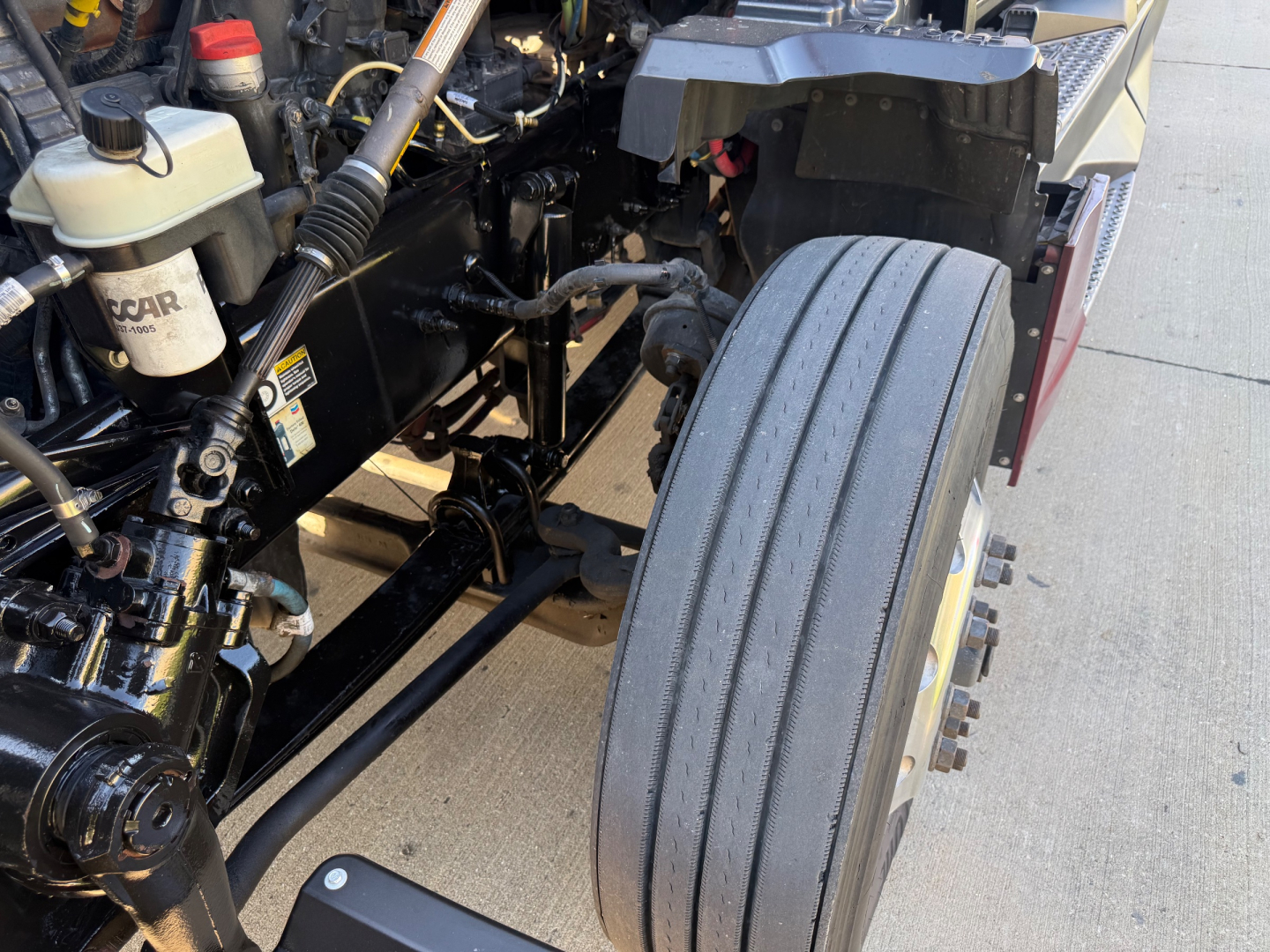 Kenworth T680 engine bay showing front axle and tire with various fluid reservoirs and hoses visible.