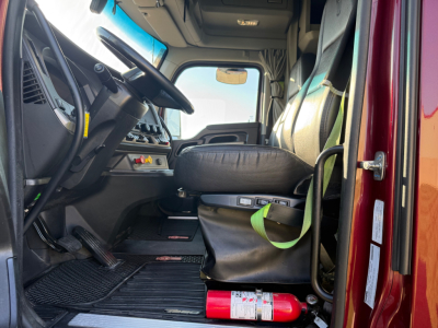 Cab interior of a 2023 Kenworth T680 with steering wheel, driver seat, and dashboard; green seat belt visible; red exterior door frame; small fire extinguisher in cab.