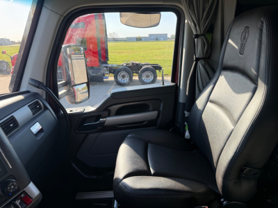 Kenworth T680 truck cab interior showing driver seat and door panel with window area; outside visible are side mirror, field, and other trucks.