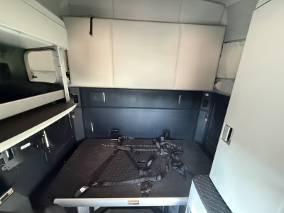 Interior of a 2023 Kenworth T680 sleeper cab with cargo area and storage racks visible, showing seating restraints and side cabinetry.