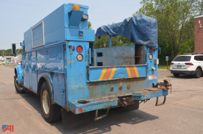 Rear and left-side angle of a blue 2002 International 4700 utility body truck with crane, showing rear platform, hoist area, and ladder access, with safety lights and reflective chevrons.