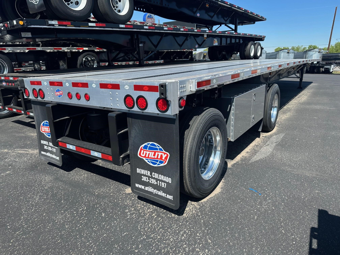 Flatbed trailer from rear-left view showing Utility spread air combo, metal deck, dual rear axles, round taillights, and branding plate mounted on mudflap.