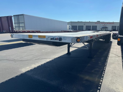 Flatbed trailer with a low-profile deck and dual rear axles, side rails, and landing gear extended. Visible are tail lights, reflectors, and a rear bumper with traction.