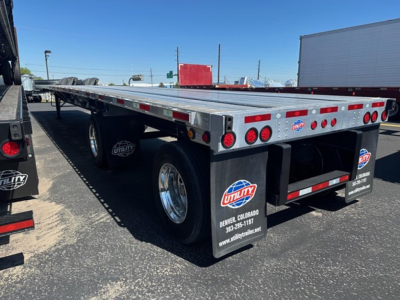 Flatbed trailer with dual wheels and rear lights, Utility branding mud flaps, silver aluminum deck, rear bumper, and reflectors; storage area and wheel well visible.
