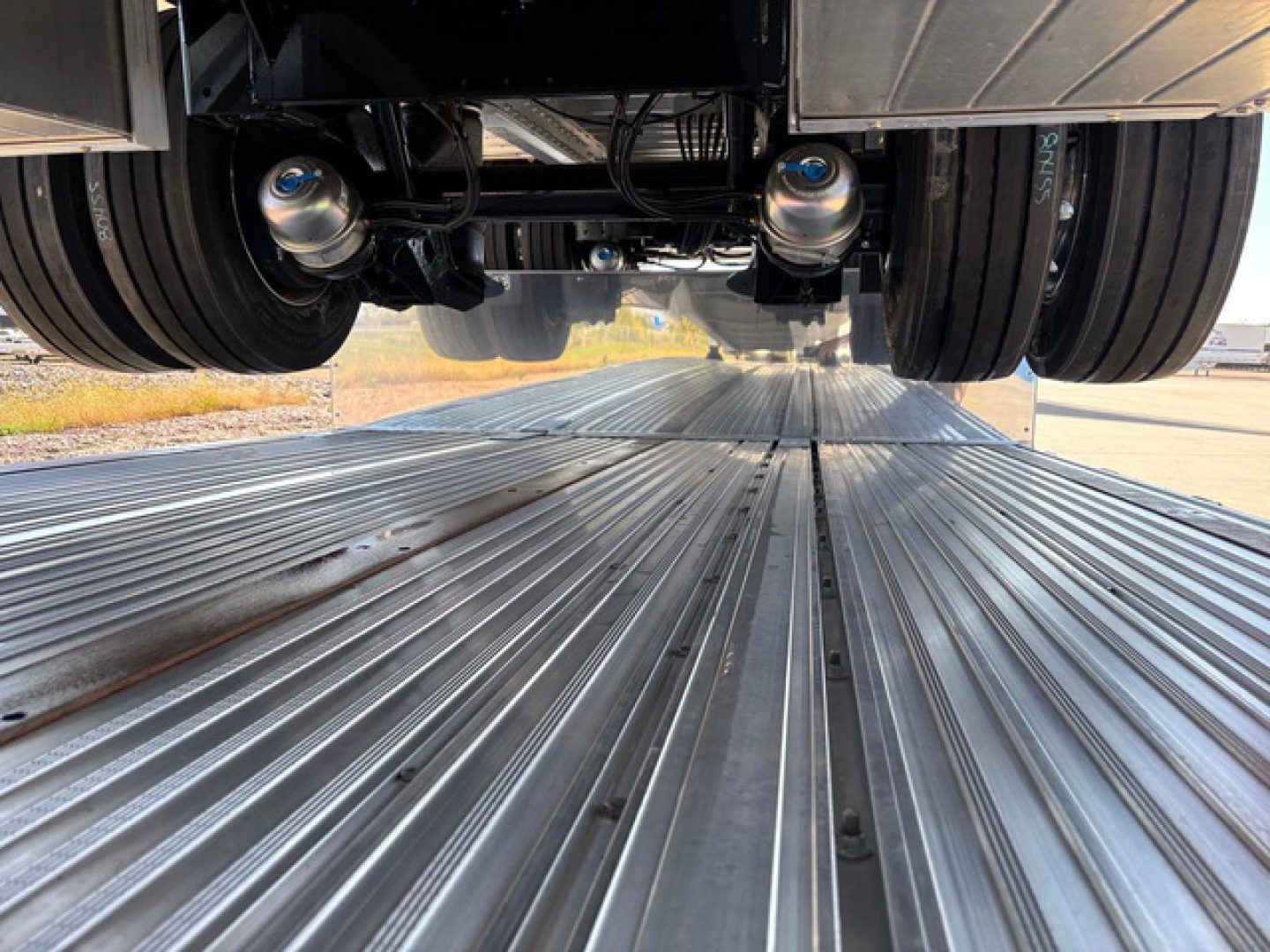 Dropdeck trailer underside with twin axles and metal deck planks, view from between the wheels looking forward