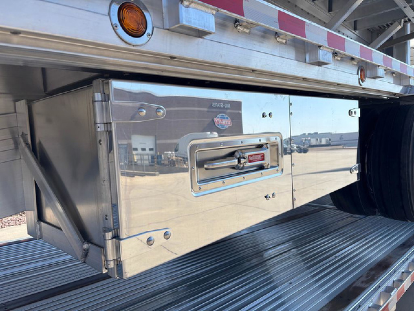 Dropdeck trailer tool chest mounted under trailer with reflective stainless surface and pins, showing storage box and wheel behind.