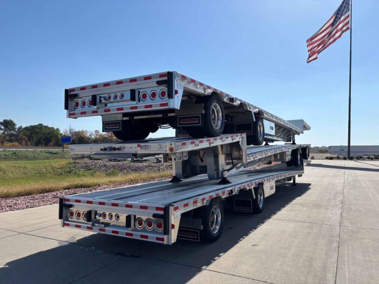 Trailer: stacked double-deck dropdeck trailer showing aluminum platform, multiple axles, lights, and American flag in background.