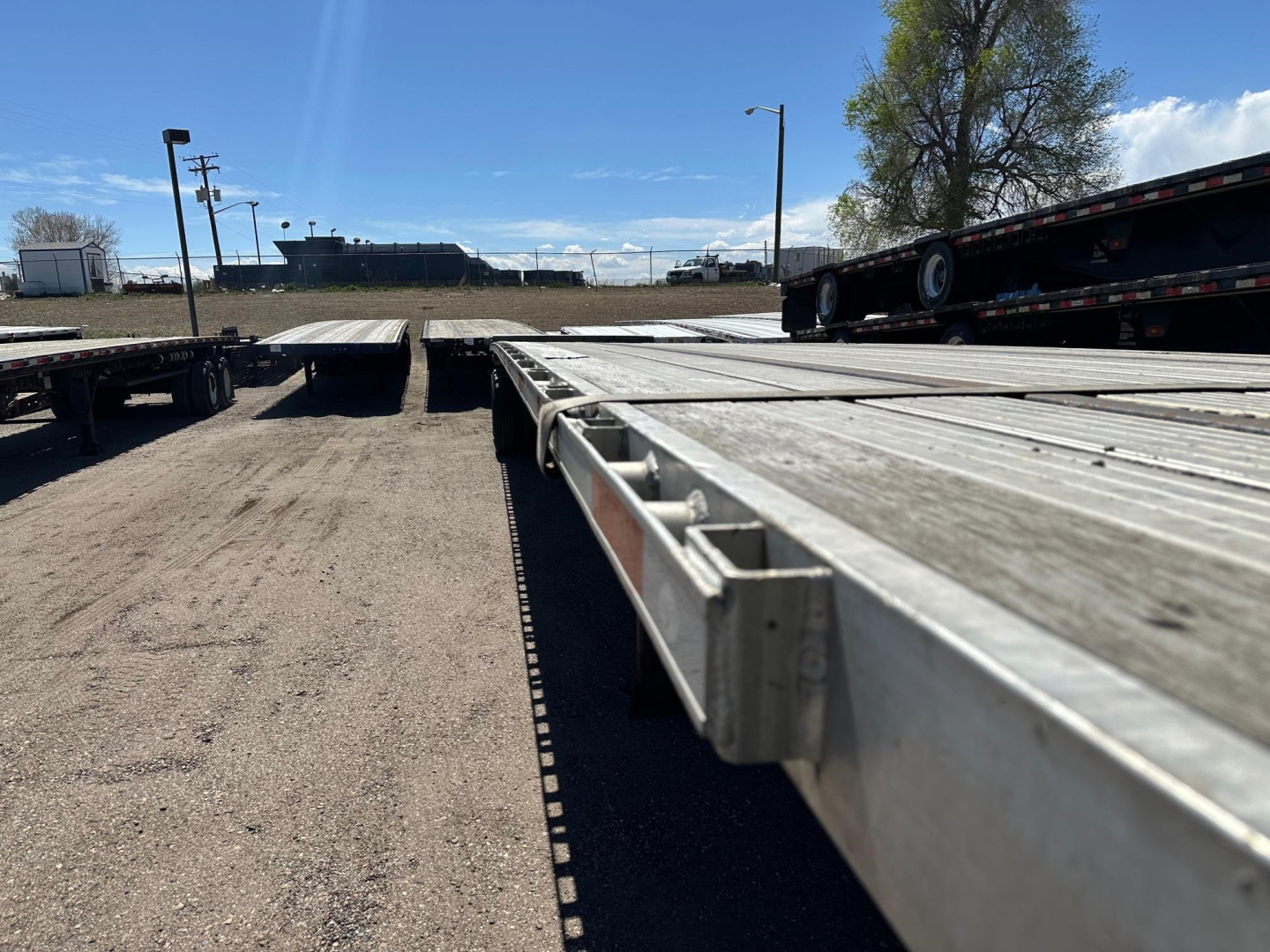 Flatbed trailer bed with metal surfaces and side rails; distant industrial yard and other trailers in background.