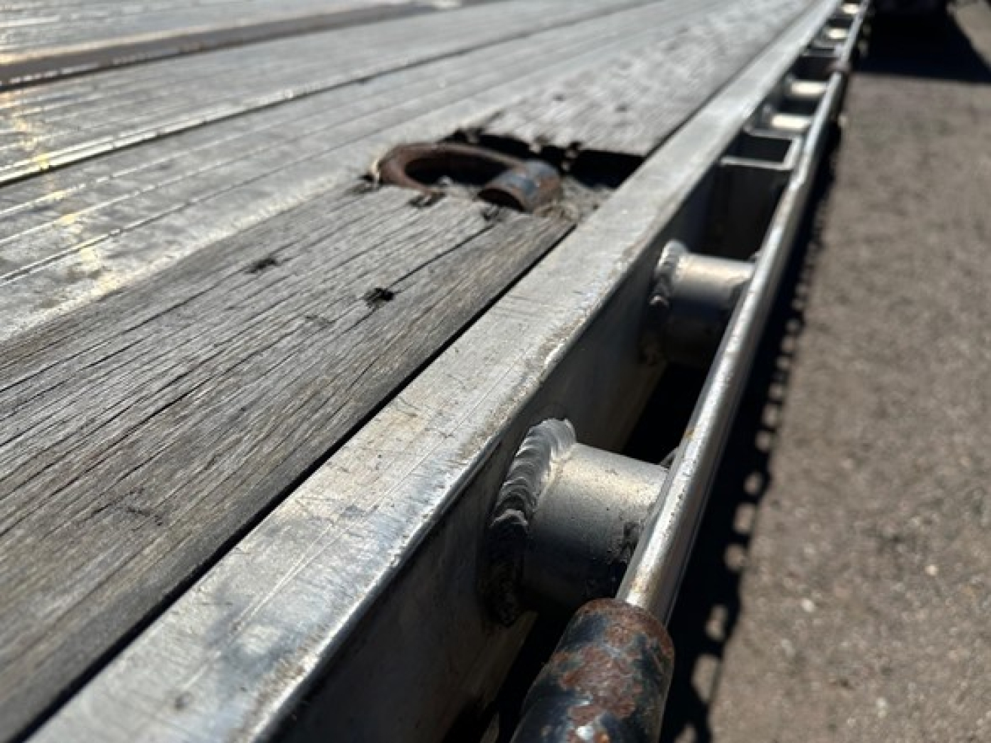 Flatbed trailer deck with metal side railing and tiedown anchors, showing wooden planks, metal edge and a rusted ring hitch area.