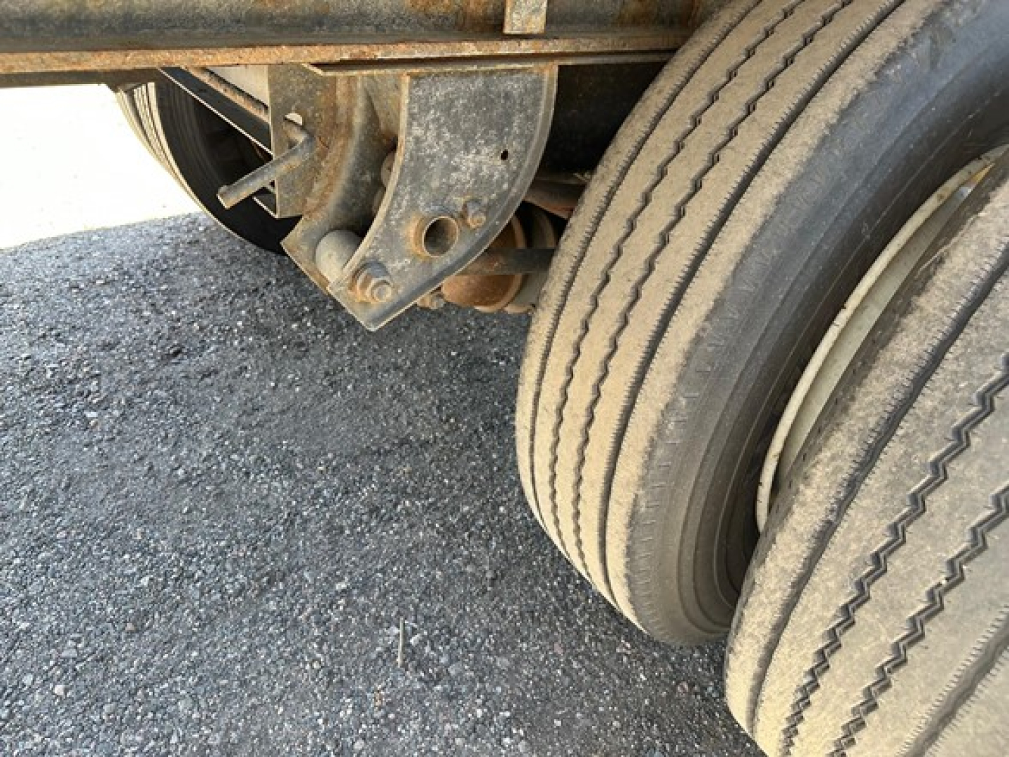 Trailer axle and dual tires under a flatbed trailer showing wheel and suspension components with visible rust and wear.