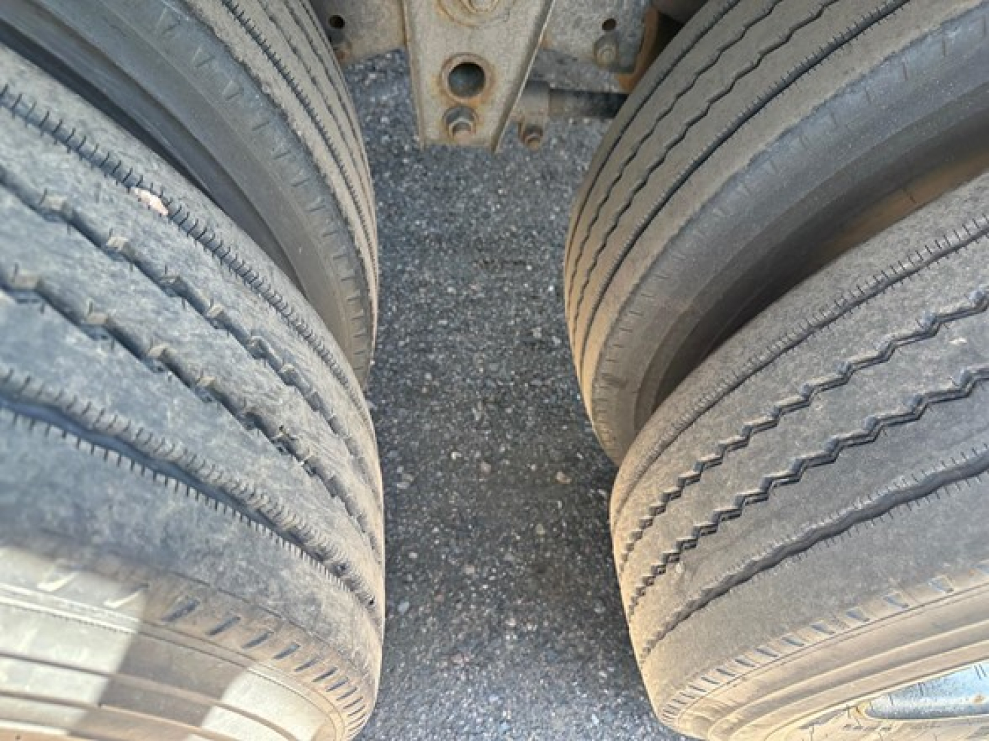 Trailer dual rear wheels and frame showing tire treads and leaf spring mounting with gravel surface.