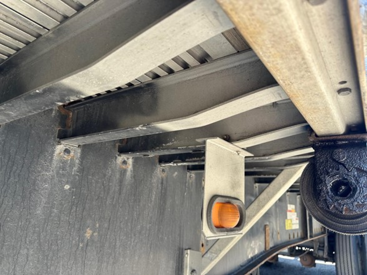Suspended underside of a utility trailer showing metal frame cross members and a cast wheel assembly with an orange knob on a silver support bracket.