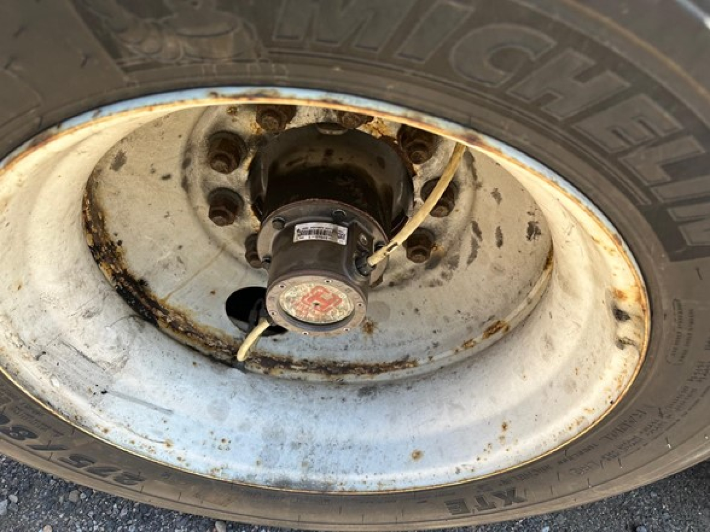 Trailer wheel hub and tire showing a steel rim with rusted areas and a central hub cap. Visible tire sidewall reads Michelin. Shows wheel and lug nuts with safety cap and a strap/line nearby.