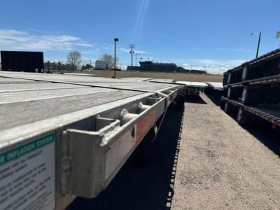 Flatbed trailer deck with stacked trailers, edge of metal frame showing a small green label; distant buildings and clear sky in background.