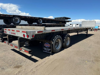 Flatbed trailer with dual rear axles and a loading ramp, Utility trailer line; spare wheel visible under deck, clear blue sky.
