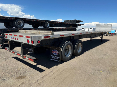 Flatbed trailer with dual rear axles and lowboy deck, Utility brand marker, showing aluminum siding and tail lights; stacks of equipment on bed.