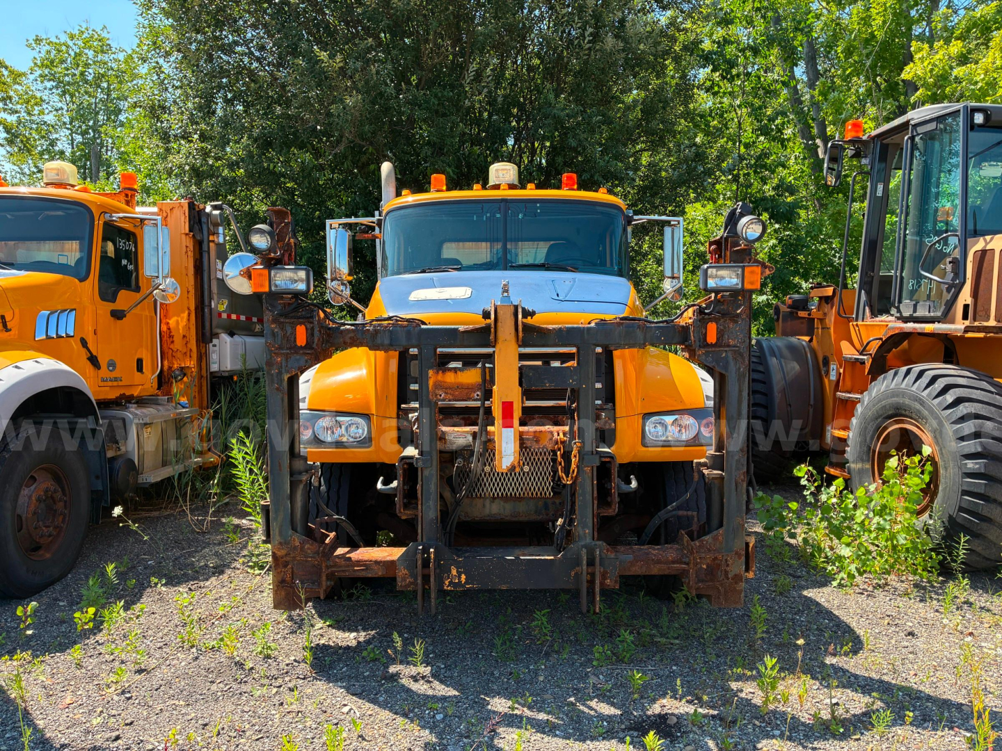Front view of a 2013 Mack GU713 dump truck with orange cab and front attachment arms, flanked by other orange trucks; trees in background.