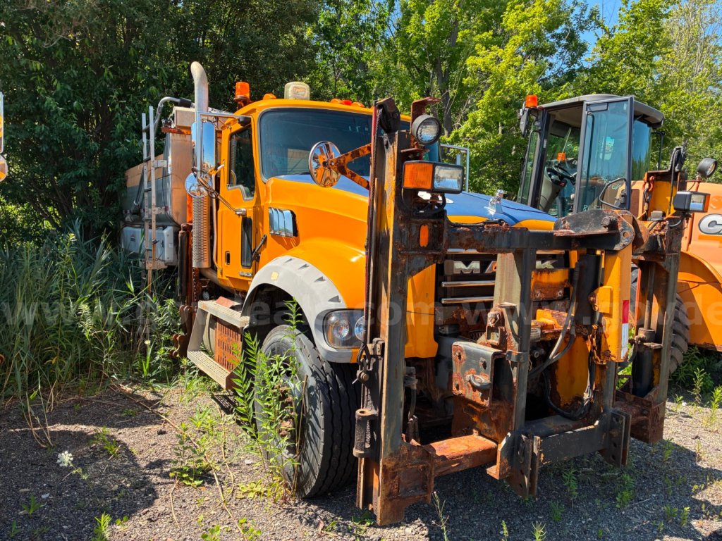 Orange 2013 Mack GU713 dump truck showing front and attachable forks in foreground, vegetation at roadside.