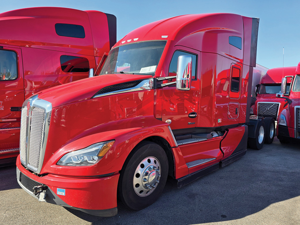 Kenworth T680 Next Gen sleeper tractor in red, side view with chrome front grille, large fuel tanks, and stacked trailers in background.