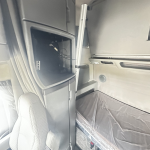 Volvo VNL860 cabin interior showing open storage compartment and vertical grab handle, sleeper area with plastic covering on mattress, door panel and window.