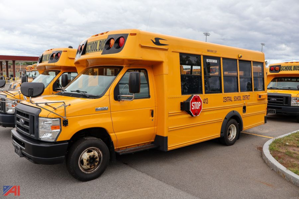 Yellow school bus van rider side view with stop sign, front grille, and Ford chassis in a bus lot.
