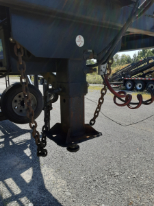Trailer hitch area showing landing gear and chains on a car carrier trailer, with a blue wheel well and a circular sticker on the upright post.