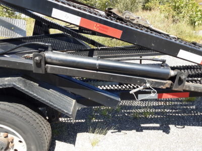 Car carrier trailer with hydraulic cylinder and deck ramps showing frame rails and wheel on left; utility chains and reflective tape visible.