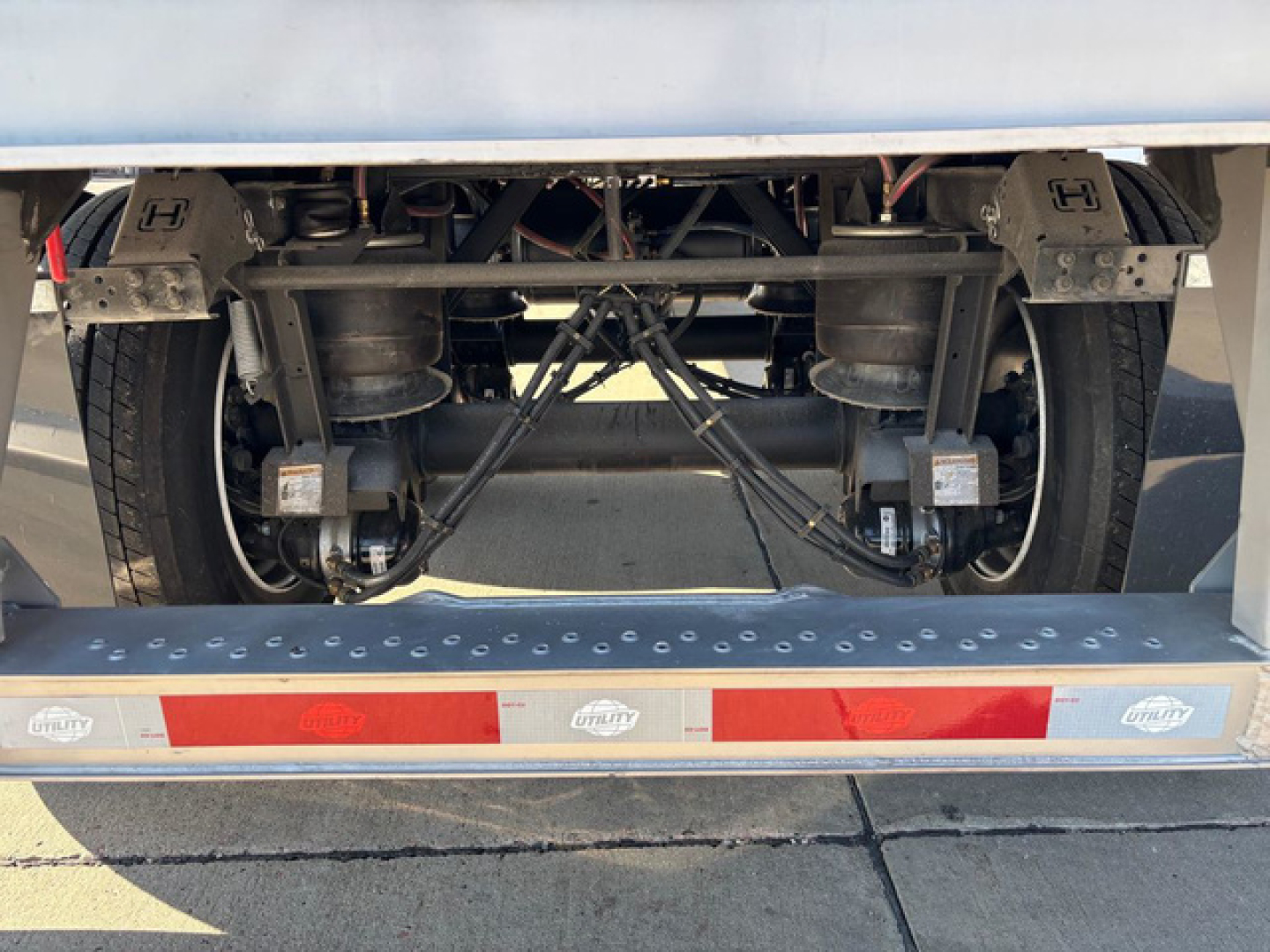 Rear axle view of a utility trailer showing dual axles, suspension components, tires, and the undercarriage with a metal step bumper and reflective red/white stripes.
