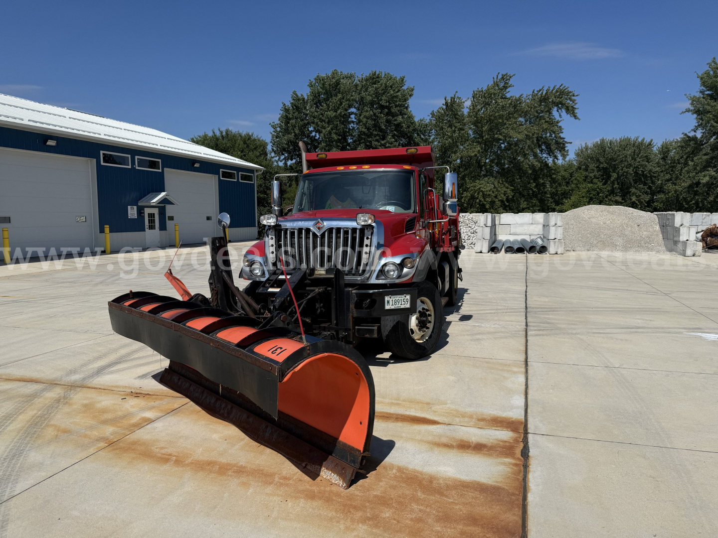 Truck and large snow plow blade attachment in front of warehouse, red 2012 International 7400 dump truck, clear day, storage yard.
