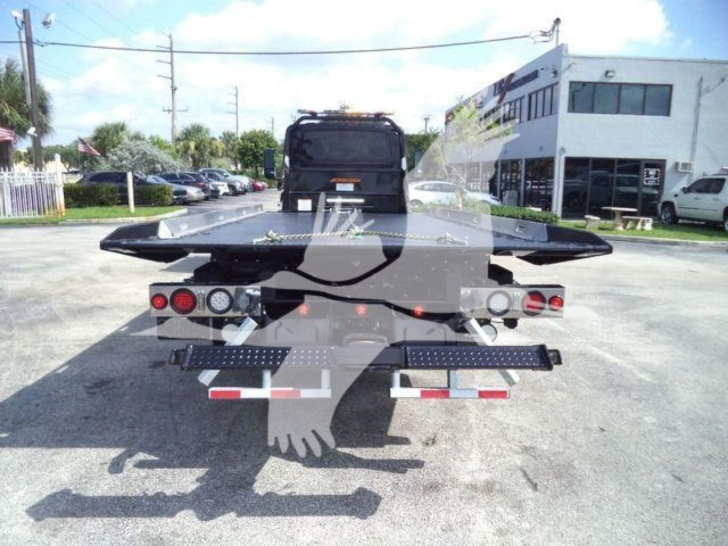 Rear view of a flatbed tow truck with platform completed by two taillights, rear bumper, and reflective tape on the sides; appears in a dealership lot.