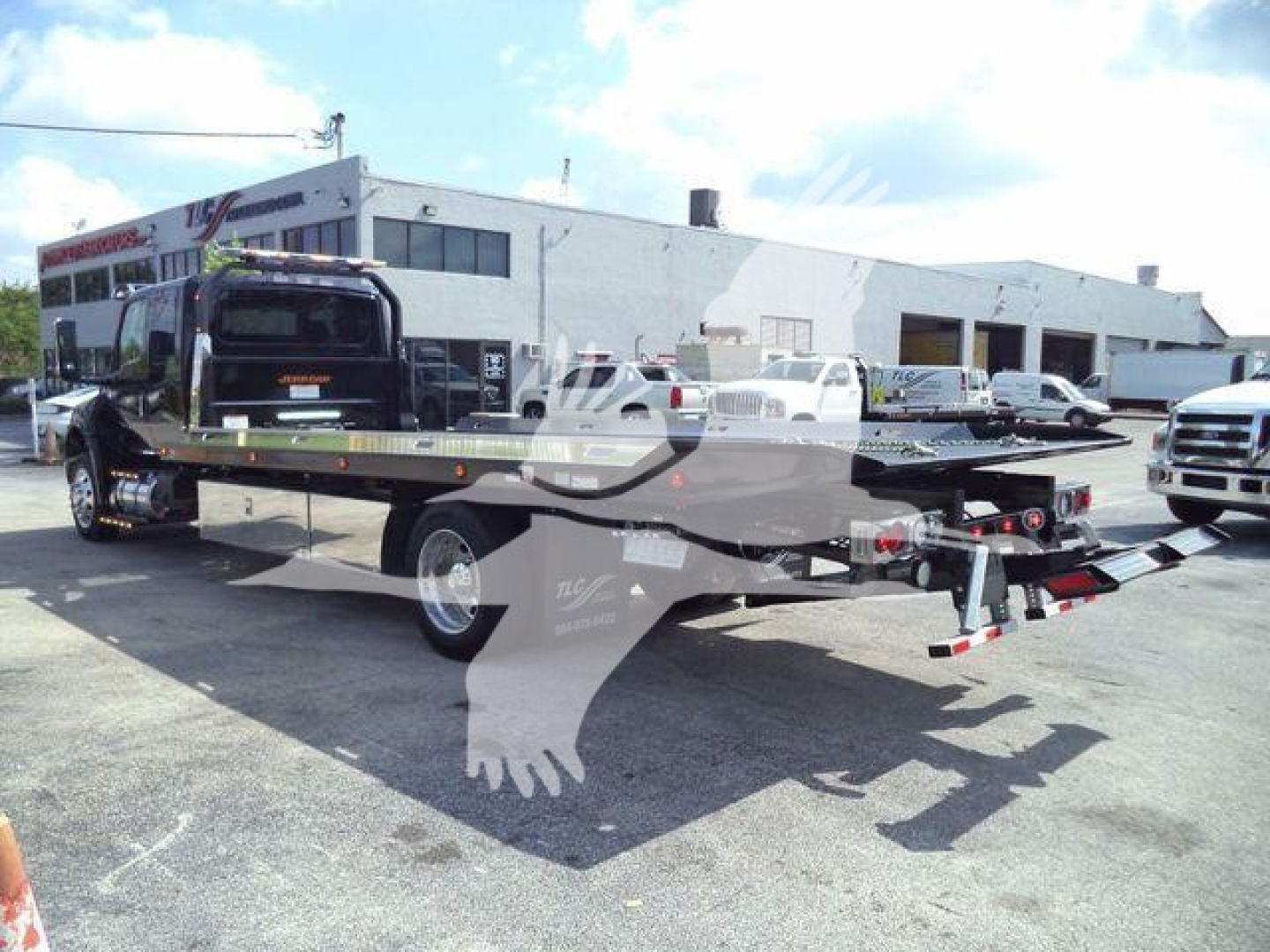 Flatbed rollback tow truck on lot with white building in background showing dual rear ramps and hydraulic hitch.
