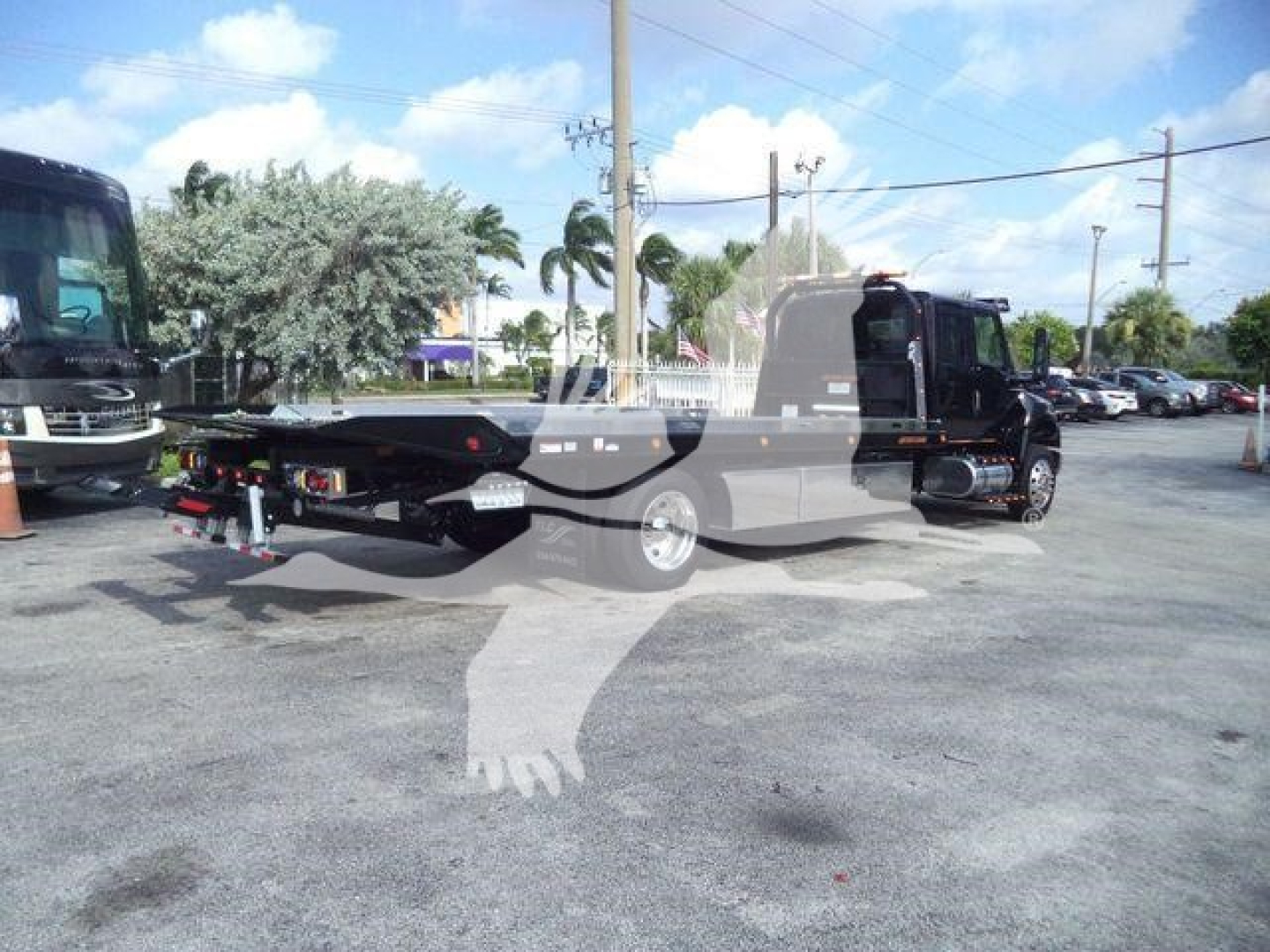 Flatbed tow truck on lot with trees and other vehicles; sticker text on side and rear not legible; watermark silhouette