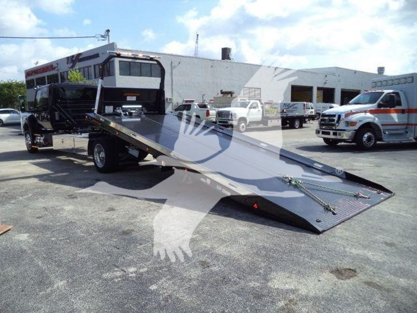 Tow truck rollback bed extended with International MV cab and flatbed ramp visible in front, service yard in background.