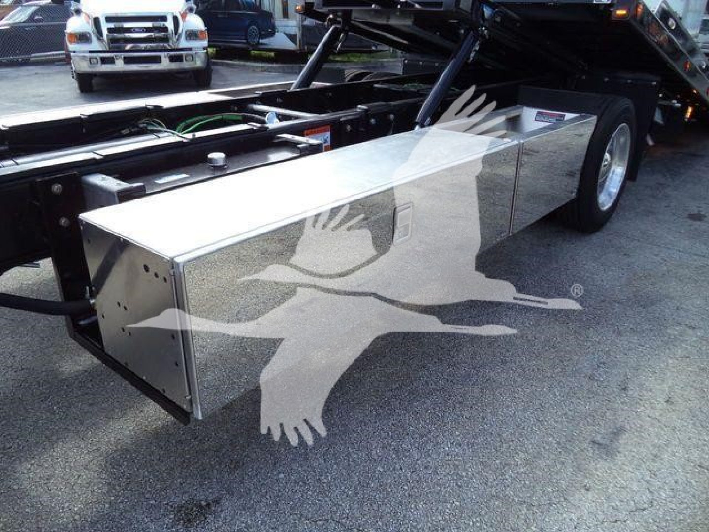 Truck rollback chassis with a large rectangular aluminum toolbox mounted beside the rear wheel. Visible components include hydraulic lines, frame rails, and a tilted rear bed with a black undercarriage.