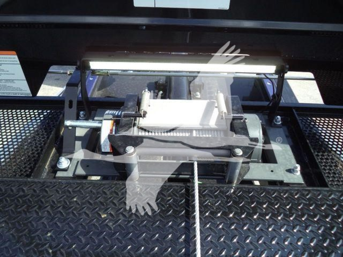 Tractor-style winch or hoist mount on a black diamond-plate surface; visible components include a silver winch drum, lever, and mounting brackets, with a white label in the background partially legible.
