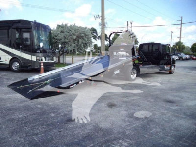 Truck transport vehicle with a large flatbed rollback ramp in a parking lot, International MV model in foreground with a tow truck and a coach bus in background.