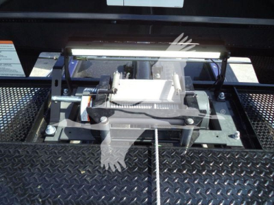 Tractor-style winch or hoist mount on a black diamond-plate surface; visible components include a silver winch drum, lever, and mounting brackets, with a white label in the background partially legible.